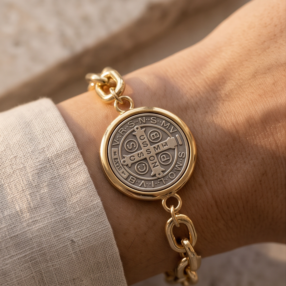 A close-up of a gold chain bracelet with a round, multi-toned medallion featuring the monogram 'CSMP' and religious text, worn on a wrist.